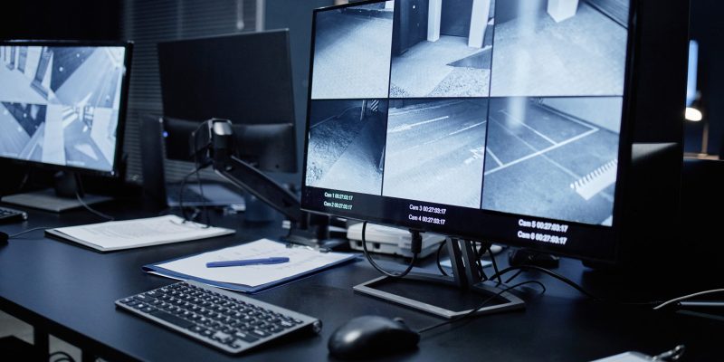 Empty security control room showing modern surveillance workstation with multiple monitors displaying live CCTV footage of building corridors and parking areas, keyboard and documents on desk