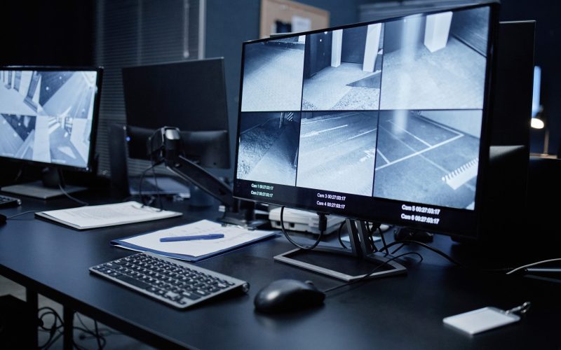 Empty security control room showing modern surveillance workstation with multiple monitors displaying live CCTV footage of building corridors and parking areas, keyboard and documents on desk